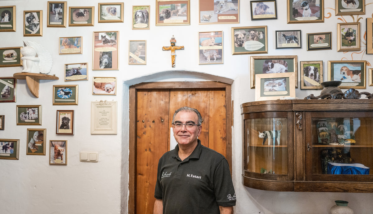 CLOUD7_ Ein Mann steht lächelnd vor einer Holztür, umgeben von einer Wand, die mit gerahmten Fotos von Haustieren bedeckt ist, und einem hölzernen Kruzifix über der Tür. Rechts davon steht eine Vitrine mit Glasfront.