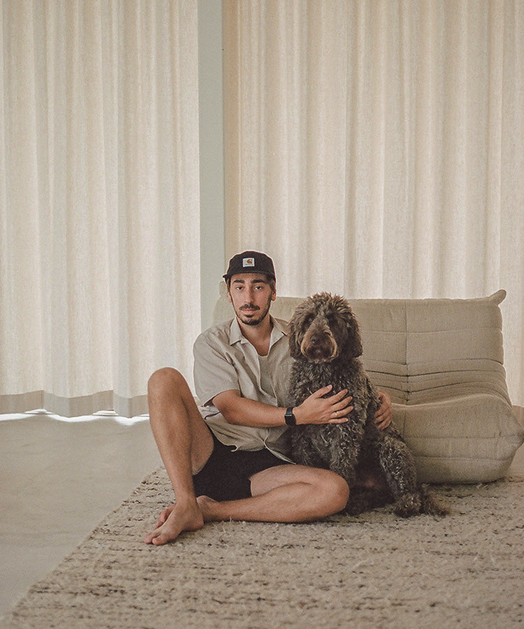CLOUD7_ Ein Mann mit einer Mütze sitzt barfuß auf einem Teppichboden neben einem flauschigen Hund. Er hat einen Arm um den Hund gelegt und schaut in die Kamera. Hinter ihnen stehen ein beiges Sofa und helle Vorhänge.