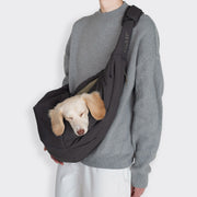 CLOUD7_ Eine Person in grauem Pullover und weißer Hose trägt einen hellen Hund in der Hundeumhängetasche LTTL Puffer Dark Grey/Khaki über der Brust. Der kuschelige Hund stützt seinen Kopf mit geschlossenen Augen auf den Taschenrand.