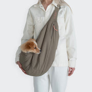 CLOUD7_ Eine Person in cremefarbenem Outfit trägt einen kleinen braun-weißen Hund in der Hundeumhängetasche LTTL Puffer Khaki/Peach über der Brust. Der Kopf des Hundes lugt seitlich vor einem schlichten weißen Hintergrund hervor.
