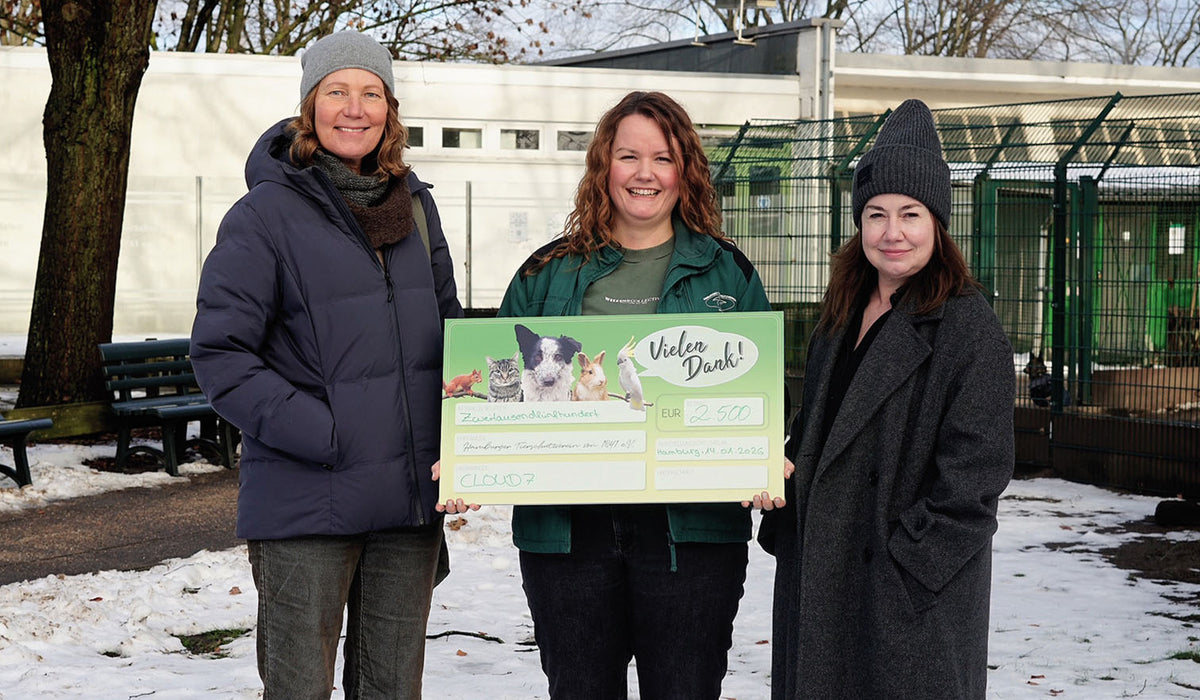 CLOUD7_ Drei Frauen stehen draußen in Winterkleidung, lächeln und halten einen großen Spendenscheck über 2.500 Euro in den Händen. Im Hintergrund sind Schnee und Bäume in der Nähe eines umzäunten Bereichs zu sehen.