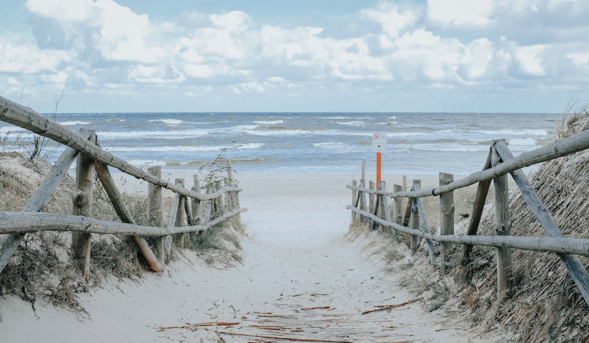 CLOUD7_ Ein sandiger Weg mit Holzgeländern führt durch die Dünen zu einem Strand mit sanften Wellen unter einem teilweise bewölkten Himmel.