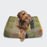CLOUD7_ Auf dem Hundebett Cube Tartan Green, einem grün-gelb-beige karierten Bett aus Bio-Baumwolle, liegt ein lockiger brauner Hund und blickt leicht zur Seite.