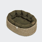 CLOUD7_ Der Hundekorb Lazy Canvas Olive ist ein kleiner, ovaler Hundekorb mit einer geflochtenen beigen Außenseite und einer gepolsterten dunkelgrünen Innenseite, die vor einem schlichten hellen Hintergrund steht.