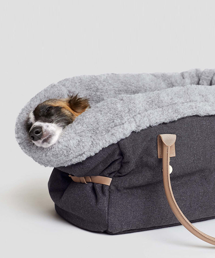 CLOUD7_ Ein kleiner Hund schläft gemütlich in einer weichen, grauen, mit flauschigem Futter ausgekleideten Transporttasche mit hellbraunen Tragegriffen und hat den Kopf auf den Rand gelegt.
