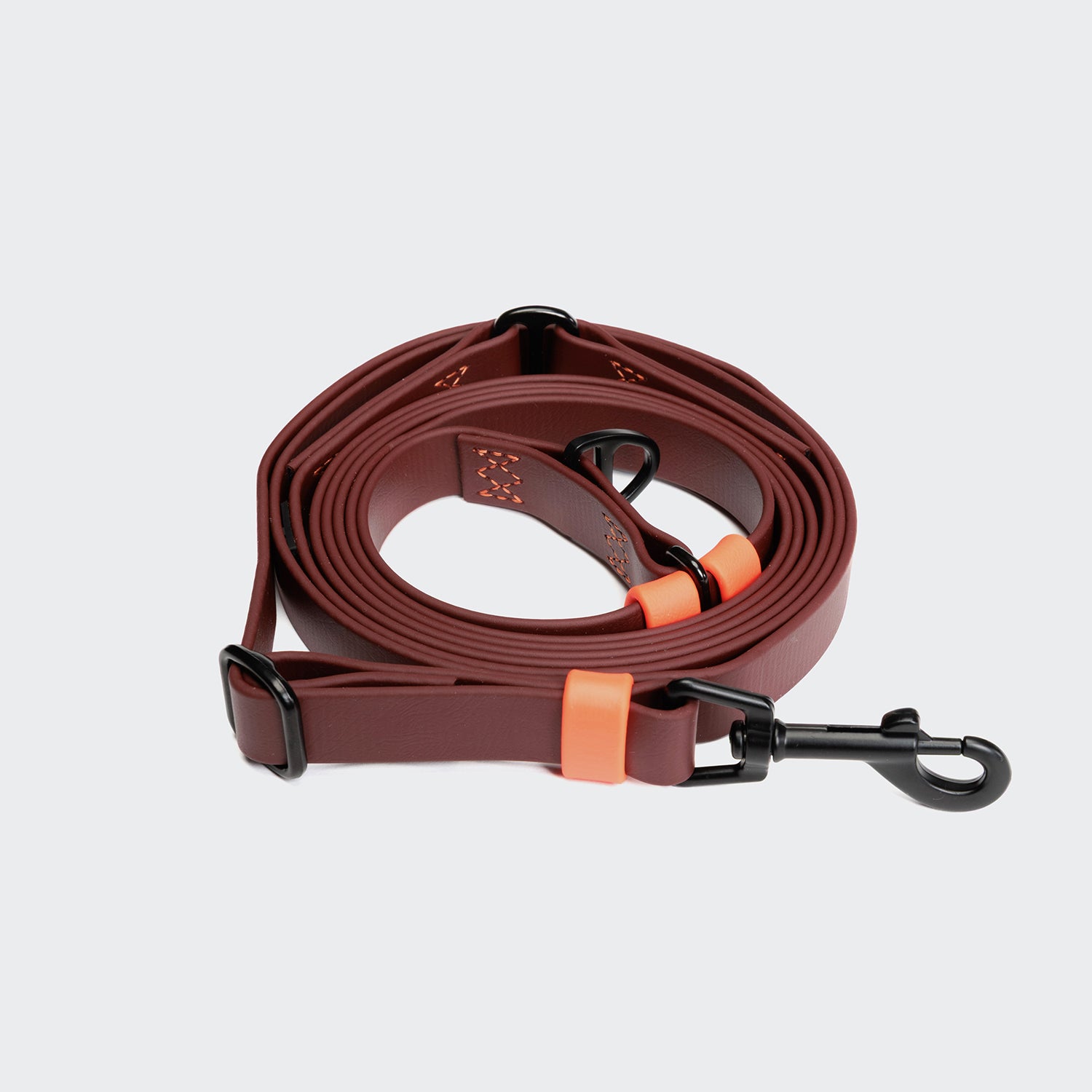 CLOUD7_ Die Freihandleine BioThane Ipanema Dark Aubergine mit orangefarbenen Akzenten und schwarzen Metallbeschlägen ist auf einem hellgrauen Hintergrund aufgerollt und dargestellt.