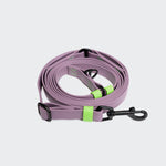 CLOUD7_ Die Freihandleine BioThane Ipanema Lilac mit grünen Akzenten und schwarzem Metallclip ist auf einem einfarbigen hellgrauen Hintergrund aufgewickelt.