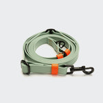CLOUD7_ Eine Freihandleine aus BioThane „Ipanema Mint-Neon Orange“ aus hellgrünem Material mit schwarzen Metallklammern und leuchtend orangefarbenen Zierstreifen ist ordentlich auf einem schlichten weißen Hintergrund aufgerollt.