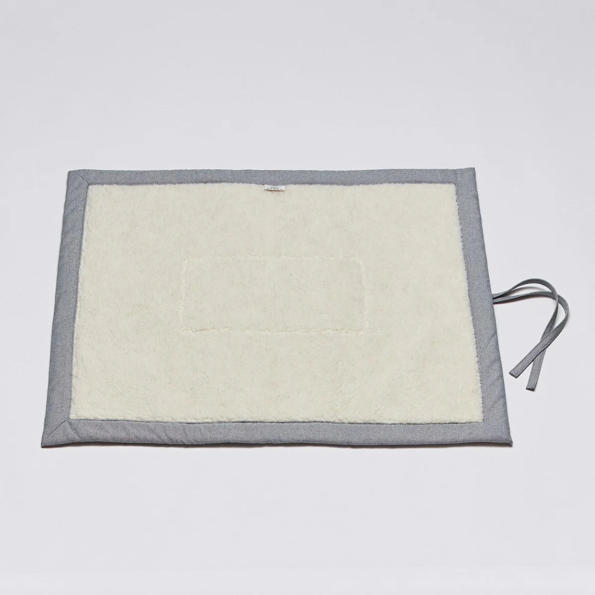 CLOUD7_ Eine rechteckige, cremefarbene Matte mit einem grauen Rand und zwei grauen Stoffbändern auf der rechten Seite, die flach auf einer schlichten weißen Fläche liegt.