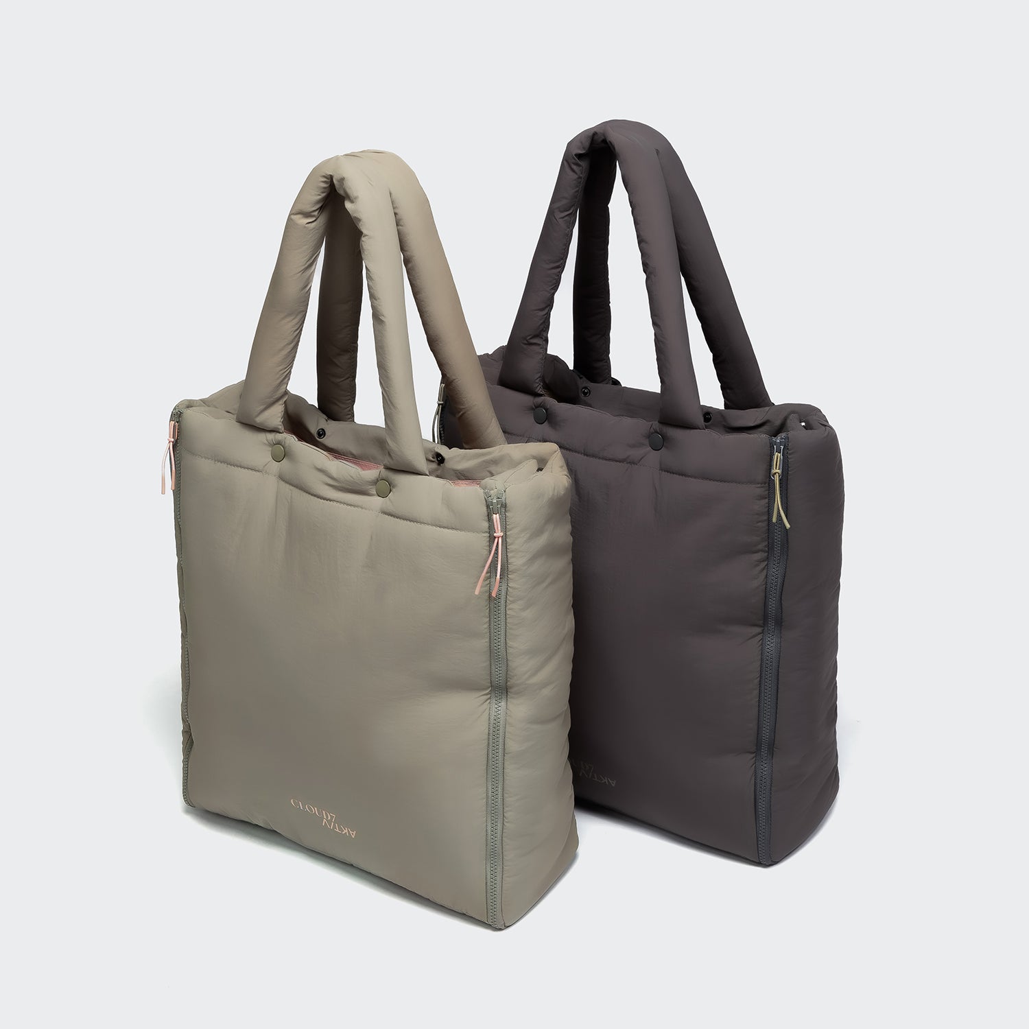 CLOUD7_ Auf einem hellgrauen Hintergrund stehen zwei gepolsterte Tragetaschen nebeneinander. Die eine ist hellbeige, die andere dunkelgrau. Beide Taschen haben kurze Henkel und einen Reißverschluss.