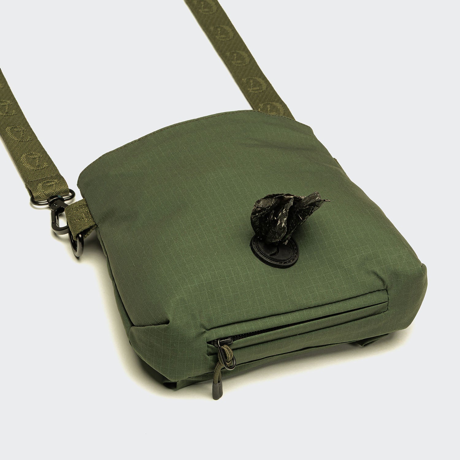 The Leckerlibeutel AKTIV Olive is a small green crossbody bag with a zippered front pocket and a roll of black waste bags partially pulled through its circular front opening. The strap has a subtle pattern—ideal for walks or Hundetraining Tasche.