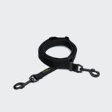 CLOUD7_ Die L-1 Freihandleine AKTIV Black ist eine aufgerollte, verstellbare Hundeleine aus recyceltem Polyester mit Metallhaken an beiden Enden, abgebildet auf einem schlichten hellgrauen Hintergrund.