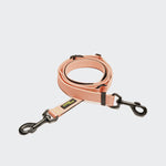 CLOUD7_ Die L-1 Freihandleine AKTIV Peach ist eine pfirsichfarbene, freihändige Hundeleine aus recyceltem Polyester mit schwarzen Metallclips an beiden Enden und einem gelb-schwarzen Markenetikett, das sauber auf einem weißen Hintergrund aufgewickelt ist.