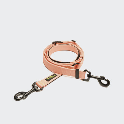 CLOUD7_ Die L-1 Freihandleine AKTIV Peach ist eine pfirsichfarbene, freihändige Hundeleine aus recyceltem Polyester mit schwarzen Metallclips an beiden Enden und einem gelb-schwarzen Markenetikett, das sauber auf einem weißen Hintergrund aufgewickelt ist.