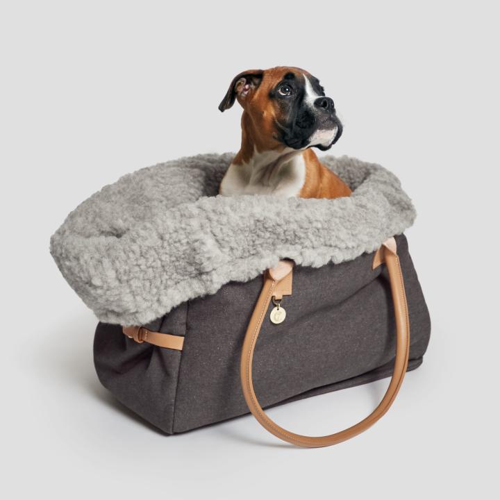 CLOUD7_ Ein braun-weißer Hund sitzt in einer weichen, grauen, mit Fleece gefütterten Tragetasche mit hellbraunen Henkeln und schaut nach oben und zur Seite. Der Hintergrund ist einfarbig hellgrau.