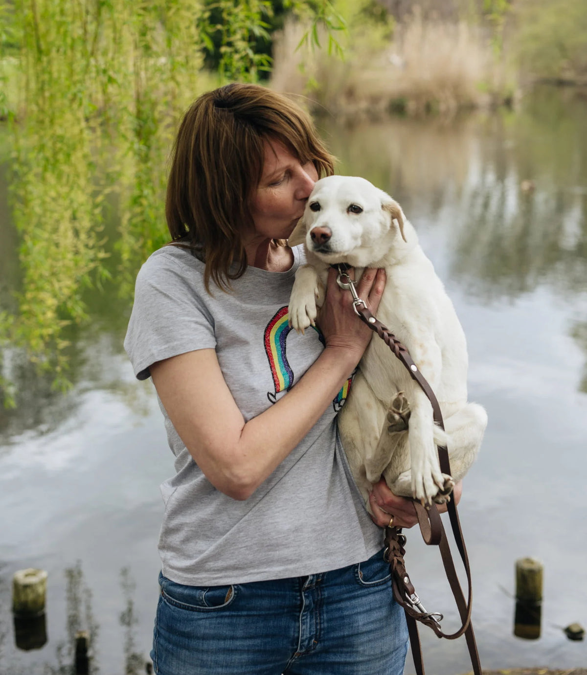 CLOUD7_ Eine Frau, die ein graues T-Shirt mit einem Regenbogenmuster trägt, umarmt und küsst sanft einen weißen Hund an einem Seeufer, umgeben von Grünflächen. Der Hund sitzt ruhig in ihren Armen und schaut in Richtung der Kamera.