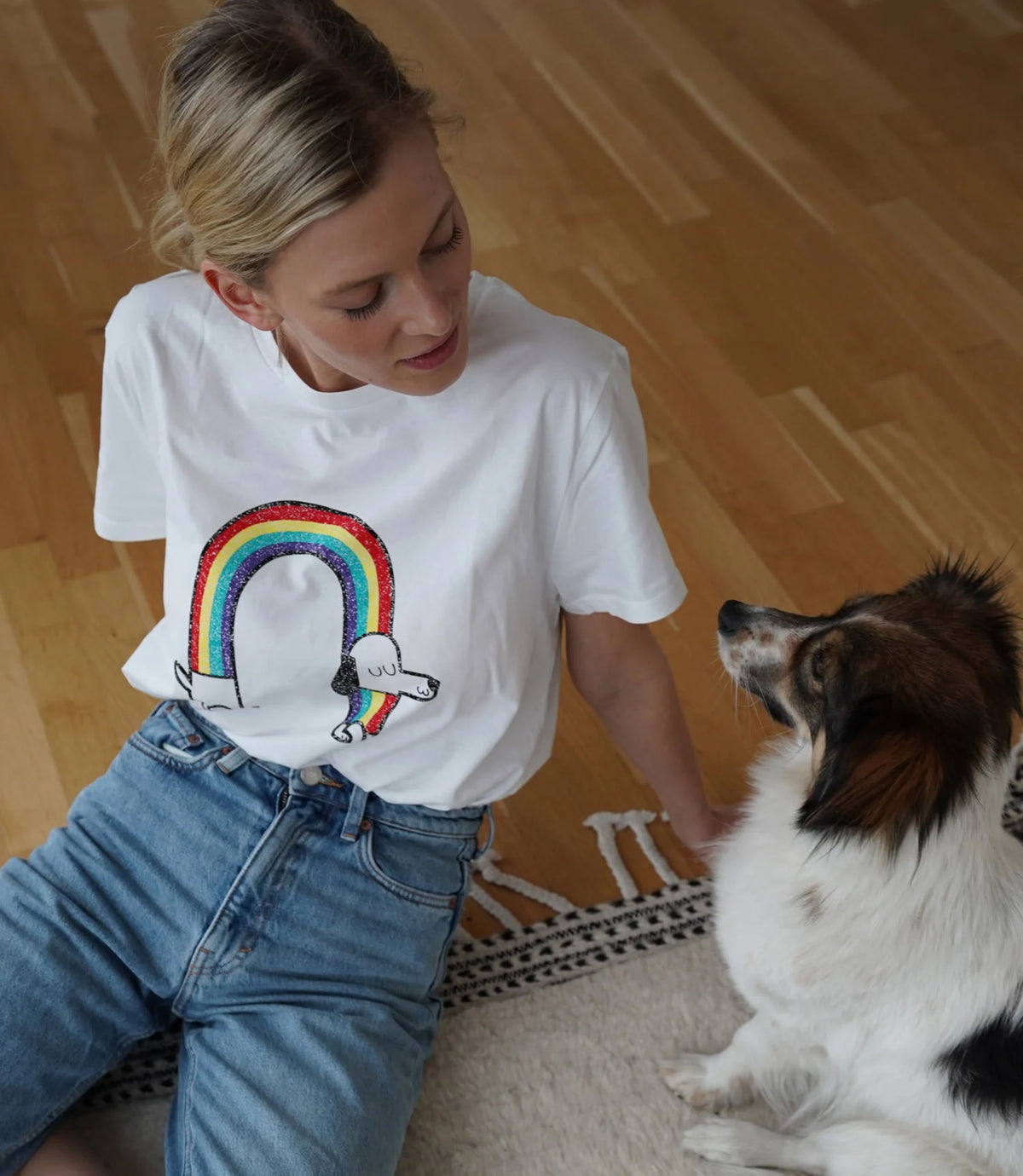 CLOUD7_ Eine Frau in einem weißen T-Shirt mit einem Regenbogen- und Dackelmotiv sitzt auf einem Holzboden und schaut einen kleinen Hund mit braun-weißem Fell an, der neben ihr sitzt.