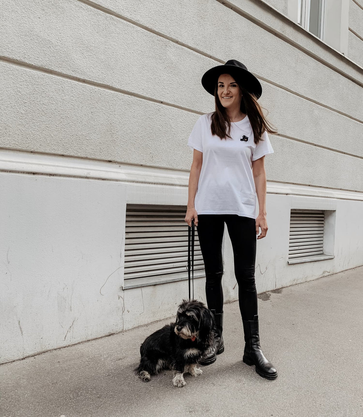 CLOUD7_ Eine Frau, die einen breitkrempigen schwarzen Hut, ein weißes T-Shirt, eine schwarze Hose und Stiefel trägt, steht auf einem Gehweg und hält einen kleinen schwarzen Hund an der Leine. Sie stehen neben einem hellen Gebäude mit belüfteten Fenstern.