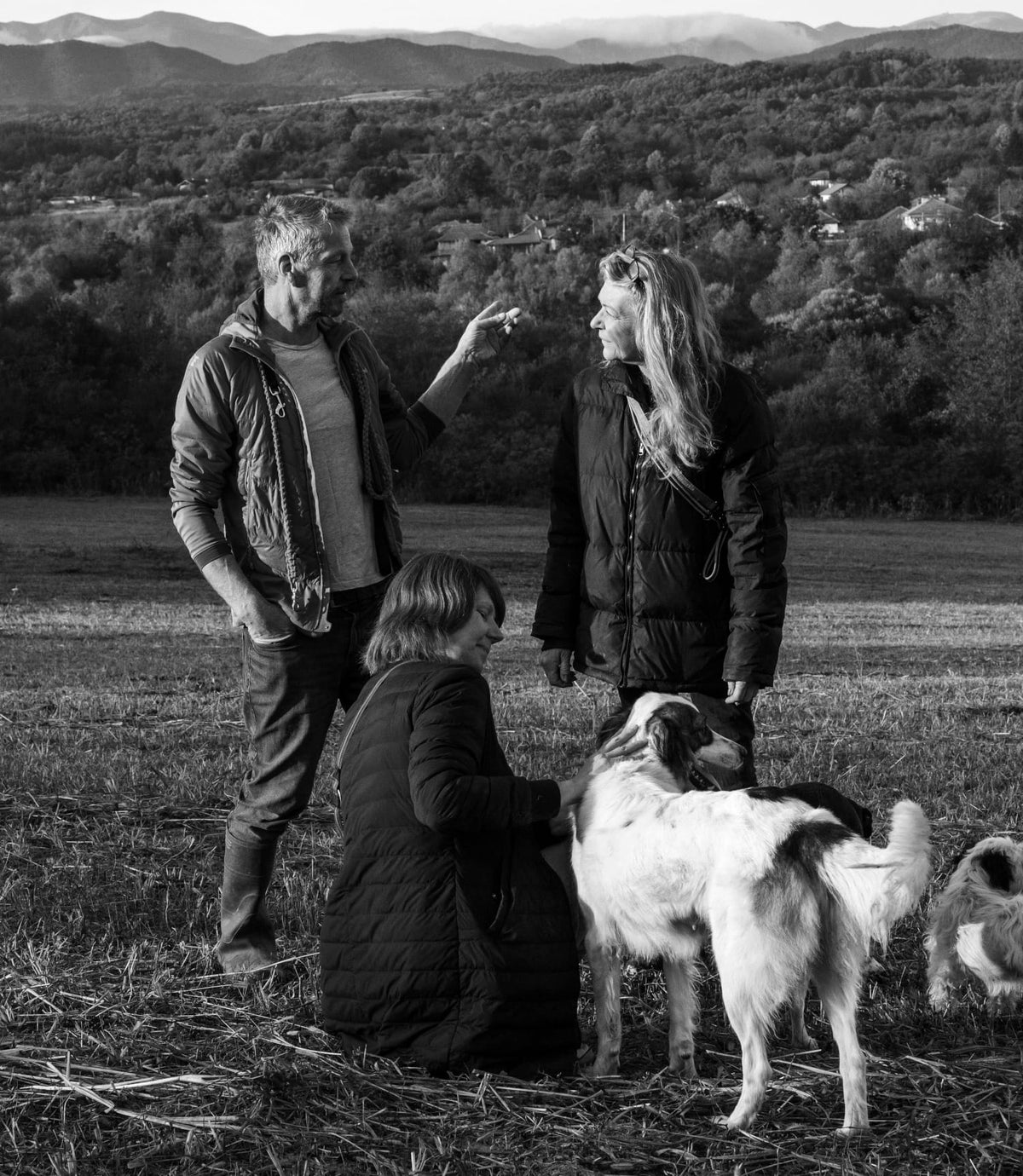 CLOUD7_ Drei Erwachsene und zwei Hunde stehen auf einem Feld, im Hintergrund sind Berge zu sehen. Eine Frau kniet nieder, um einen Hund zu streicheln, während die beiden anderen Personen stehen und sich unterhalten. Die Szene ist in Schwarz-Weiß gehalten.