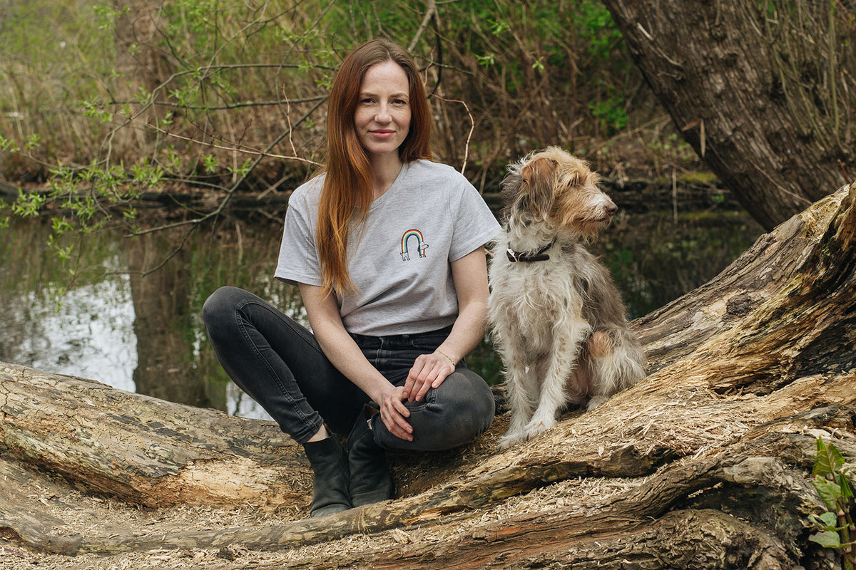 CLOUD7_ Eine Frau mit langen roten Haaren sitzt auf einem umgestürzten Baum am Ufer eines Teiches, neben ihr ein struppiger Hund. Sie trägt ein graues T-Shirt und schwarze Jeans, während der Hund zur Seite schaut. Der Hintergrund ist bewaldet und naturbelassen.