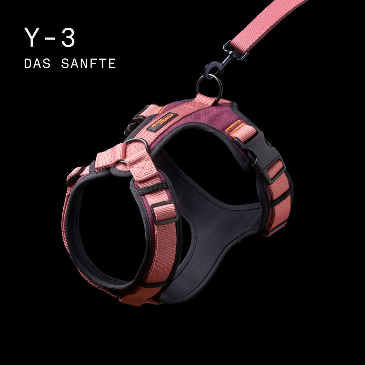 CLOUD7_ Rosa-schwarzes Hundegeschirr mit verstellbaren Riemen und einer daran befestigten Leine, dargestellt vor schwarzem Hintergrund. In der oberen linken Ecke steht in weißer Schrift „Y-3 DAS SANFTE“.
