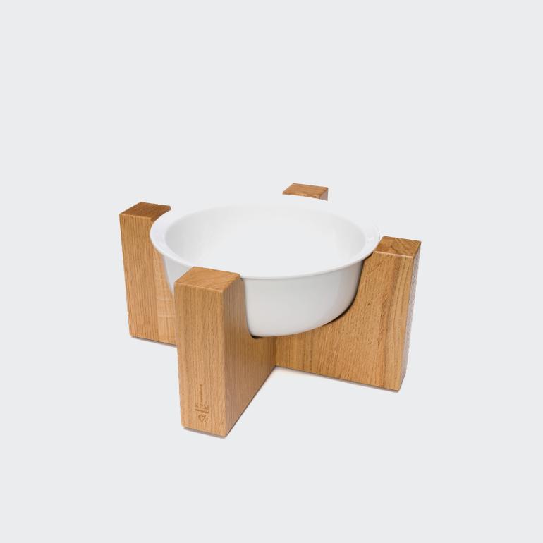 CLOUD7_ Eine weiße Keramikschale ruht auf einem Holzständer mit vier senkrechten, rechteckigen Beinen, wodurch ein modernes, minimalistisches Design entsteht. Der Hintergrund ist einfarbig und hell.
