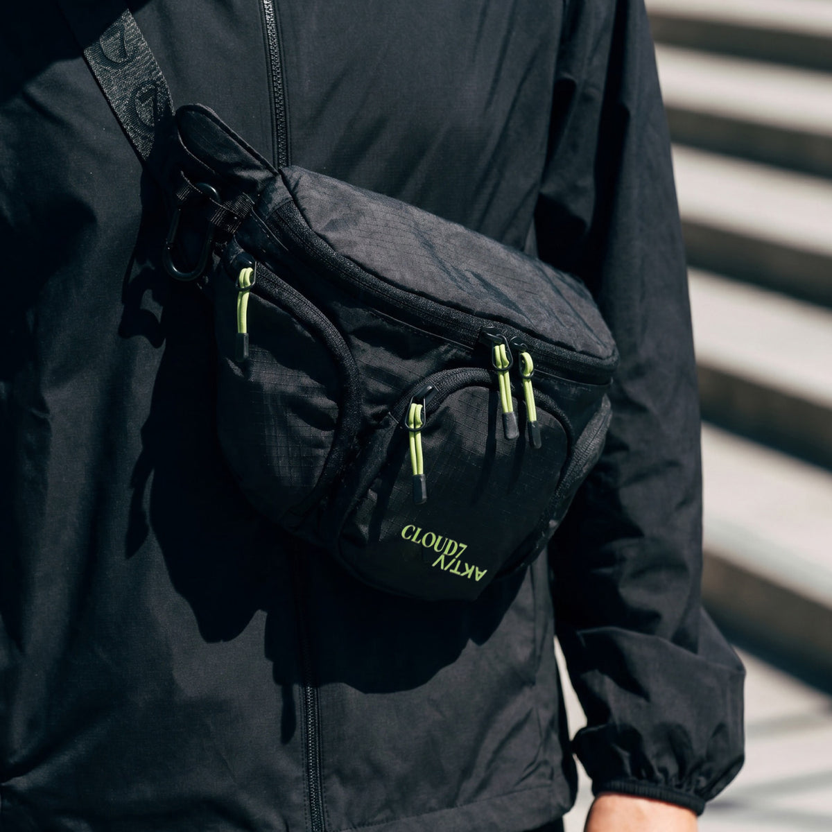CLOUD7_ Eine Person, die eine schwarze Jacke mit Reißverschluss und eine schwarze Umhängetasche mit neongrünen Reißverschlüssen trägt, auf deren Vordertasche in grüner Schrift „CLOUDY DAY“ steht.
