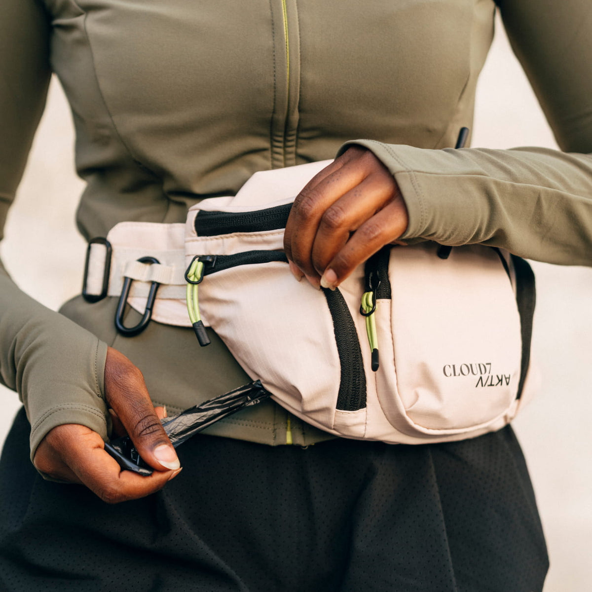 CLOUD7_ Eine Person, die eine beige Gürteltasche trägt, öffnet den Reißverschluss einer Tasche. Sie hat dunkle Haut und trägt eine olivgrüne Jacke und schwarze Shorts.