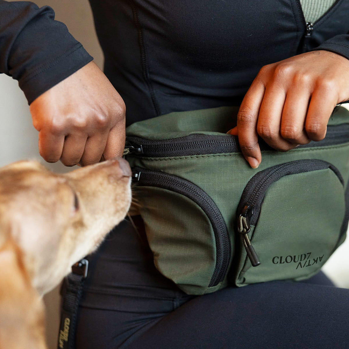 CLOUD7_ Eine Person in schwarzer Sportkleidung öffnet eine grüne Gürteltasche mit der Aufschrift „CLOUD7“, während ein brauner Hund an der Tasche schnüffelt.