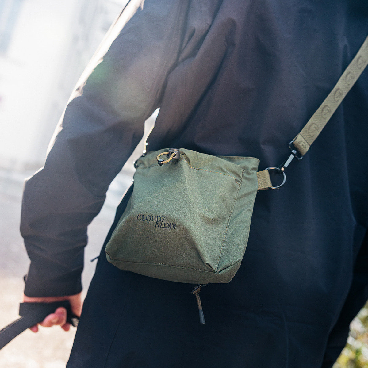 CLOUD7_ Eine Person in einer schwarzen Jacke trägt eine grüne Umhängetasche mit der Aufschrift „CLOUD7“, steht draußen im Sonnenlicht und hält in der linken Hand eine Leine oder einen Riemen.