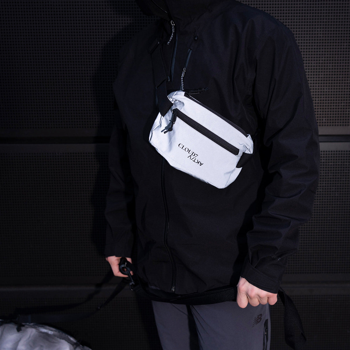 CLOUD7_ Eine Person in schwarzer Jacke und grauer Hose, mit einer weißen Umhängetasche mit der Aufschrift „CLOUD ARMY“ über der Brust, die vor einem dunklen, strukturierten Hintergrund steht.