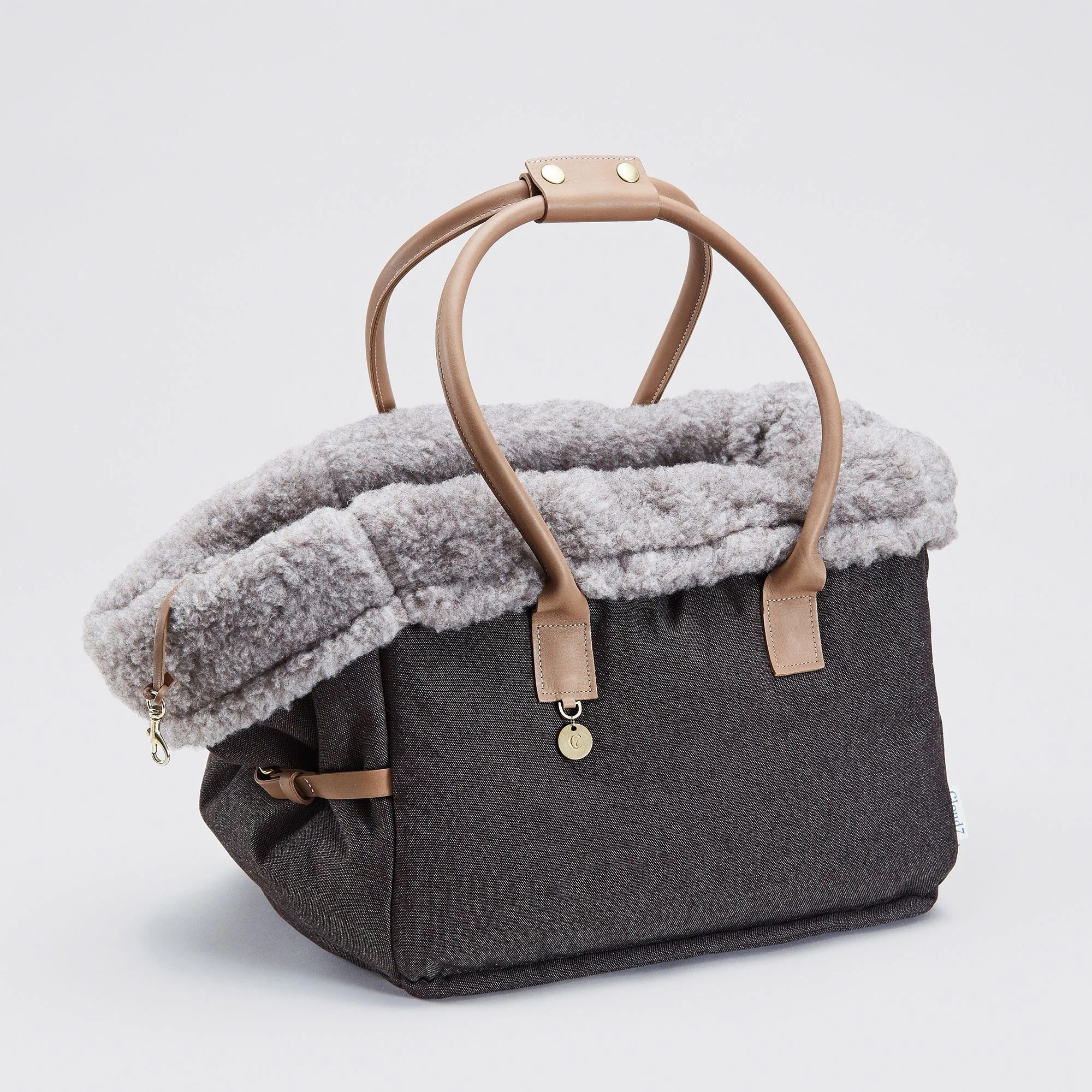 CLOUD7_ Graue Tragetasche mit hellbraunen Henkeln und Einfassungen, mit einem weichen, flauschigen grauen Futter am oberen Rand und einem goldenen Anhänger, der an einem der Henkel hängt. Die Tasche ist vor einem schlichten weißen Hintergrund abgebildet.