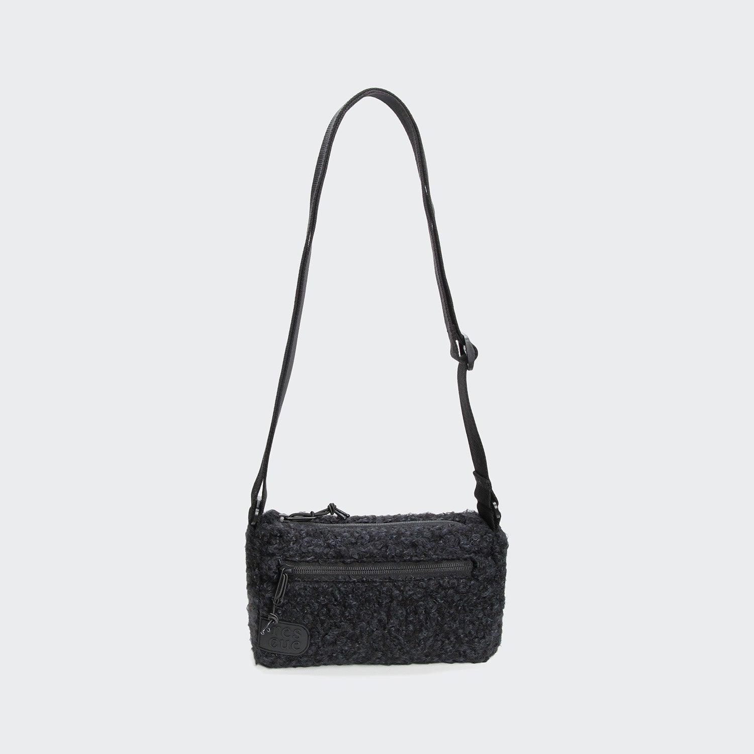 CLOUD7_ Die RESC7UE Leckerlitasche Plush Anthra ist eine kleine, rechteckige, strukturierte schwarze Crossbody-Tasche mit einem verstellbaren Riemen, einer Reißverschlusstasche auf der Vorderseite und einem Reißverschluss mit rundem Etikett, der vor einem einfarbigen hellgrauen Hintergrund abgebildet ist.
