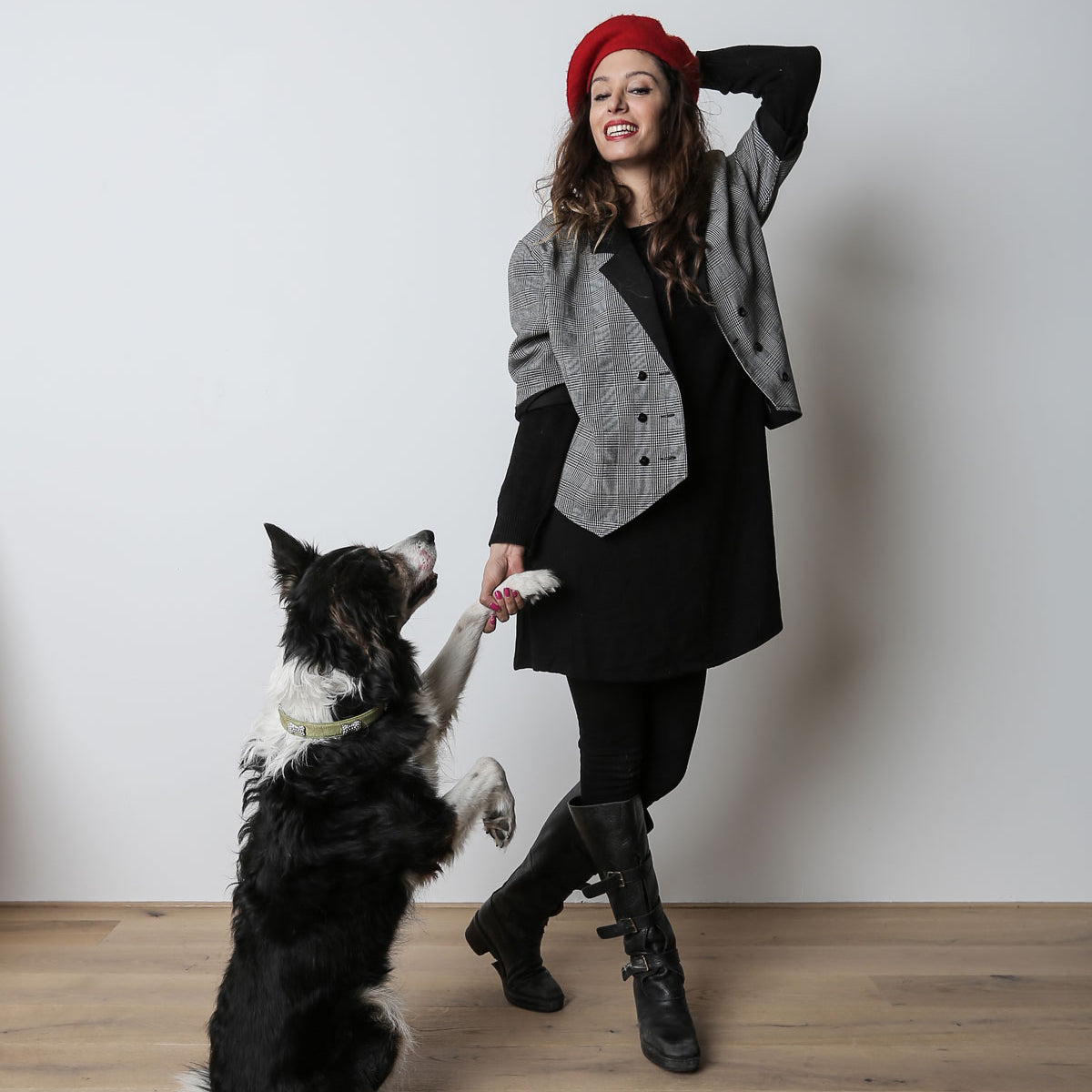 CLOUD7_ Eine Frau mit rotem Barett und grauer Jacke lächelt, während sie die Pfote eines schwarz-weißen Hundes hält. Der Hund steht auf seinen Hinterbeinen. Sie befinden sich in einem Raum auf einem Holzboden vor einer schlichten weißen Wand.