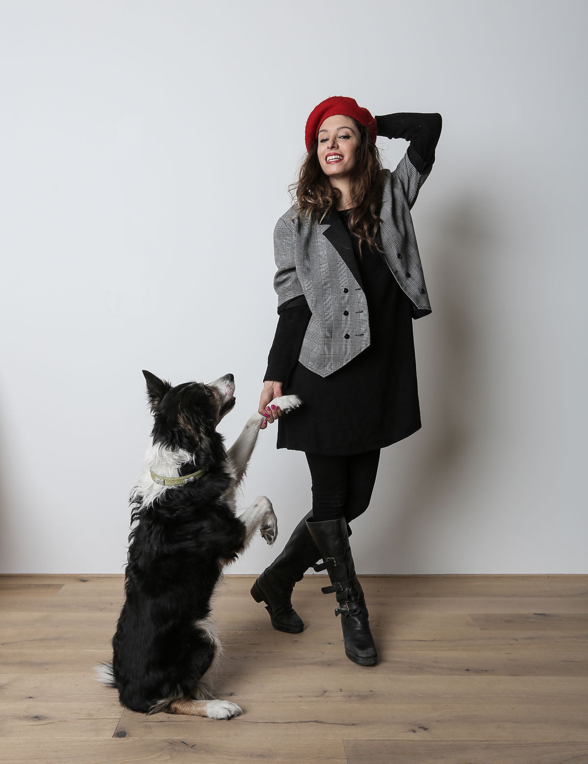 CLOUD7_ Eine Frau mit rotem Barett und grauer Jacke lächelt, während sie die Pfote eines schwarz-weißen Hundes hält. Der Hund steht auf seinen Hinterbeinen. Sie befinden sich in einem Raum auf einem Holzboden vor einer schlichten weißen Wand.