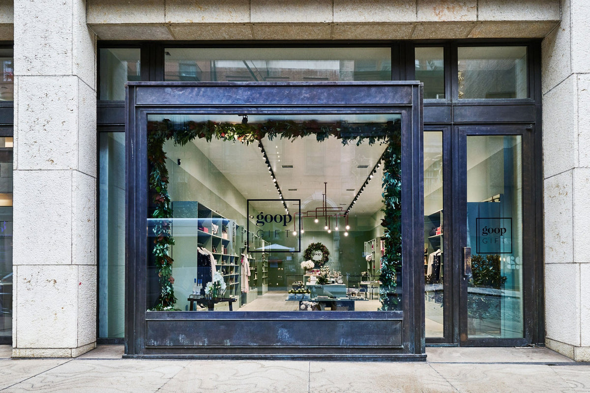 CLOUD7_ Ladenfront mit großem Schaufenster, das einen modernen, hell beleuchteten Innenraum zeigt, der mit Grünpflanzen und einem Kranz geschmückt ist. Im Inneren sind Regale mit Produkten zu sehen, und auf dem Fenster und der Tür steht „goop GIFT“.