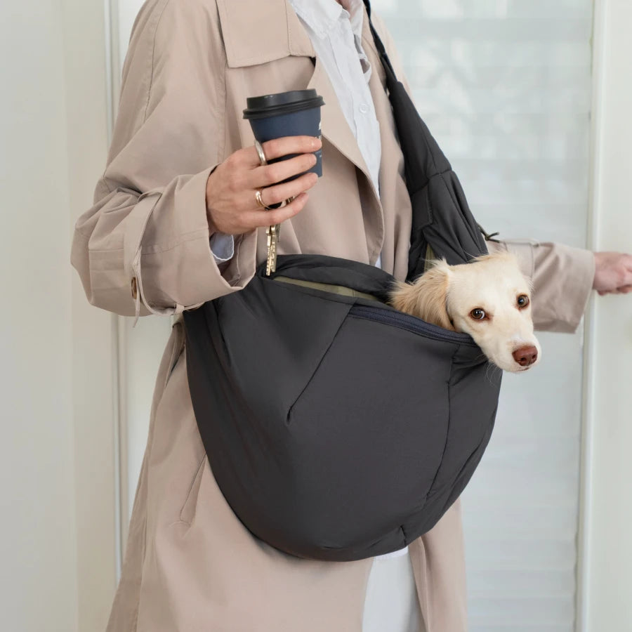 CLOUD7_ Eine Person in einem beigen Mantel hält eine Kaffeetasse in der Hand und trägt einen kleinen Hund in einer schwarzen Umhängetasche. Der Kopf des Hundes guckt heraus und schaut zur Seite.