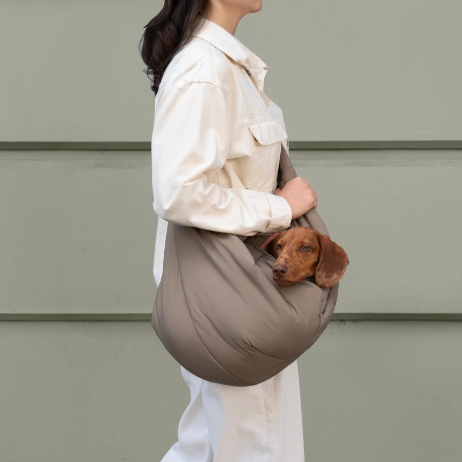 CLOUD7_ Vor einer gedeckten grünen Wand steht eine Frau in einem cremefarbenen Outfit, die einen kleinen braunen Hund in einer taupefarbenen Umhängetasche trägt.